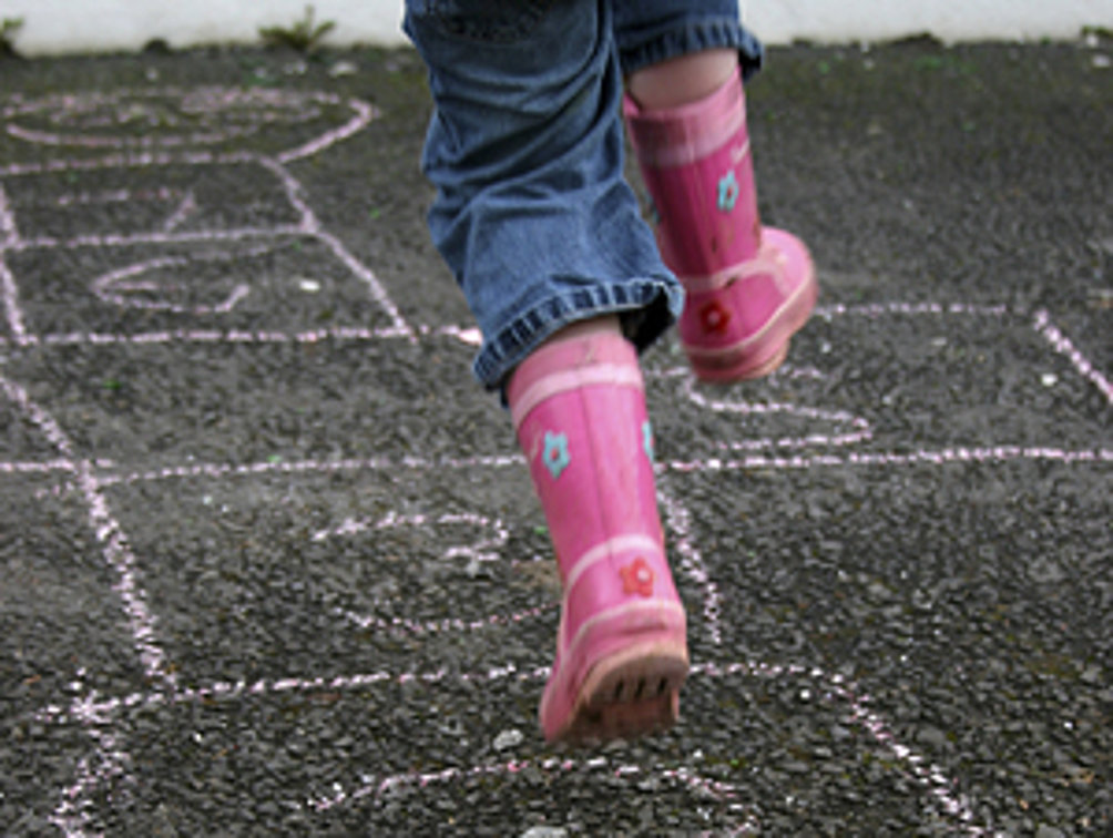 Ein Kind in rosa Gummistiefeln springt auf einem mit Kreide gezeichneten Hüpfspiel